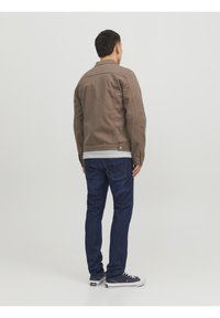 Hnědá džínová bunda s límcem a knoflíkovým zapínáním, nošená přes pruhované tričko. Spárovaná s tmavě modrými džíny a teniskami. Pohled zezadu.