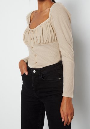 Mujer con top beige abotonado con manga larga fruncida y vaqueros negros de tiro alto con una mano en el bolsillo sobre fondo blanco.