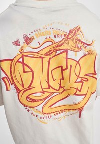 Hvid t-shirt med et stort gult og rødt graffiti-inspireret grafisk design, der viser stiliseret tekst og en slange med buede linjer og accenter.