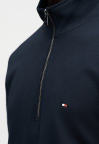 Φούτερ σε ναυτικό μπλε με φερμουάρ τεταρτοκεφαλής σε ασημένιο χρώμα και μικρό κεντητό ορθογώνιο λογότυπο σε κόκκινο, λευκό και μπλε χρώμα στο στήθος.