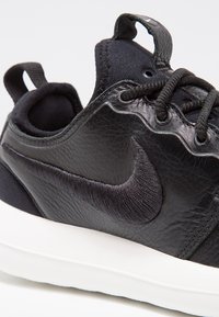 Gros plan sur une basket Nike en cuir noir avec un cuir texturé, des lacets noirs, le logo brodé et une semelle blanche.