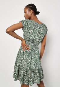 Grünes und cremefarbenes, gemustertes Kleid mit rüschensäumigem Saum, kurzen Ärmeln und einem gerafften Bund. Der Stoff hat ein strukturiertes, fließendes Aussehen.