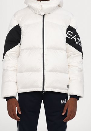 Osoba ubrana w biały puchowy płaszcz z czarnymi łatami na łokciach i czarne spodnie z białym logo EA7 na lewej udzie.