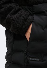 La main d'une personne dans la poche d'une veste matelassée noire avec une manche en polaire, montrant le curseur de la fermeture éclair et l'écusson avec le logo "ICEPEAK".