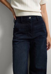 Donkerblauwe wijde jeans met een soepele textuur. Heeft een knoopsluiting en twee voorkruiszakken. Geribbelde witte top zichtbaar erboven.