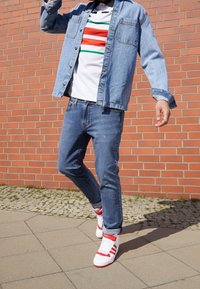 Džínová bunda přes proužkované bílé tričko s červenými, zelenými a červenými pruhy; doplněno o slim fit modré džíny a bílé tenisky s červenými detaily.