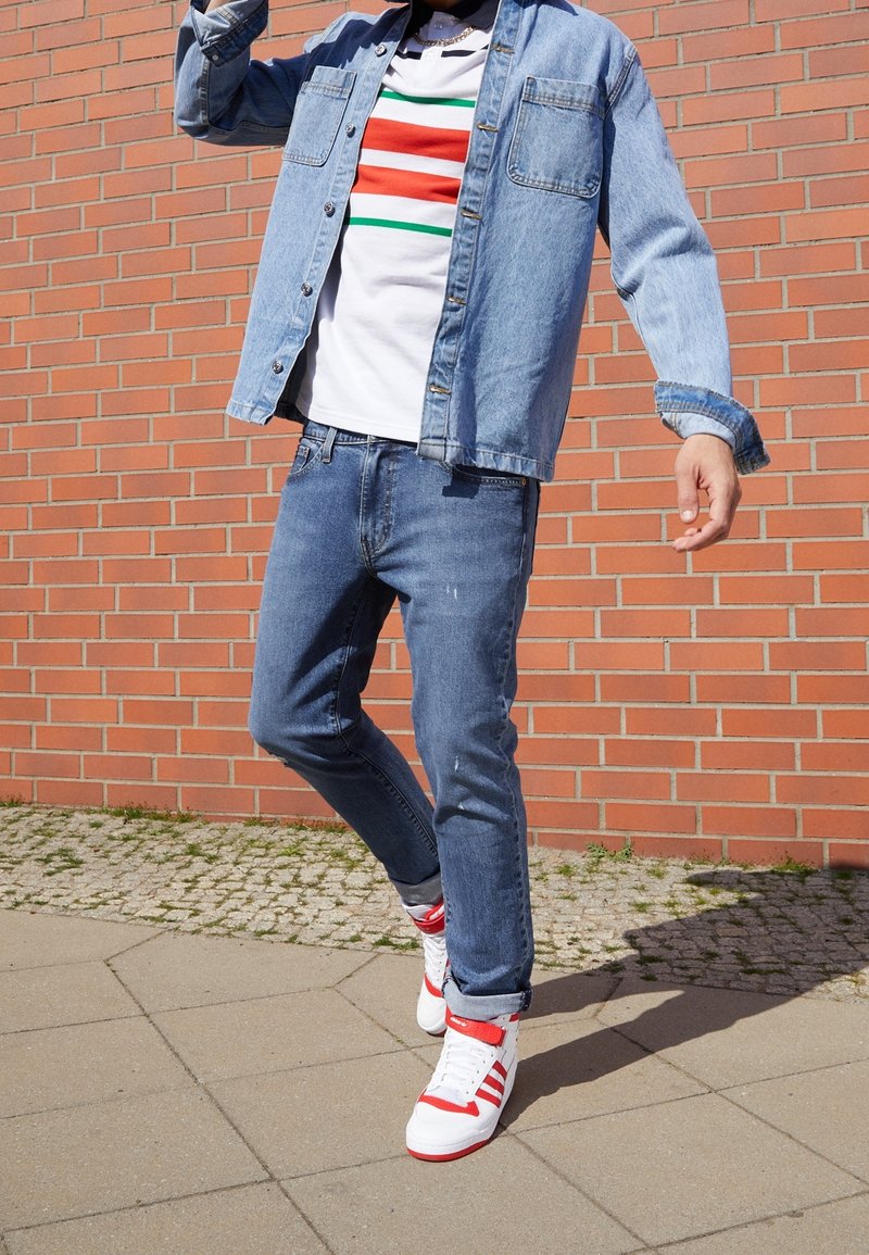 Džínová bunda přes proužkované bílé tričko s červenými, zelenými a červenými pruhy; doplněno o slim fit modré džíny a bílé tenisky s červenými detaily.