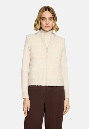 Kvinde med kort blondt hår bærer en cremefarvet, fluffy ærmeløs vest over en beige rullekrave og mørkebrune bukser mod en ensfarvet baggrund.