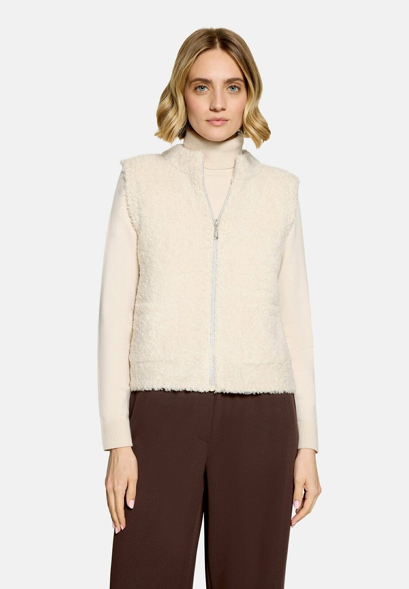 Femme aux cheveux blonds courts porte un gilet sans manches duveteux couleur crème par-dessus un col roulé beige et un pantalon marron foncé sur un fond uni.