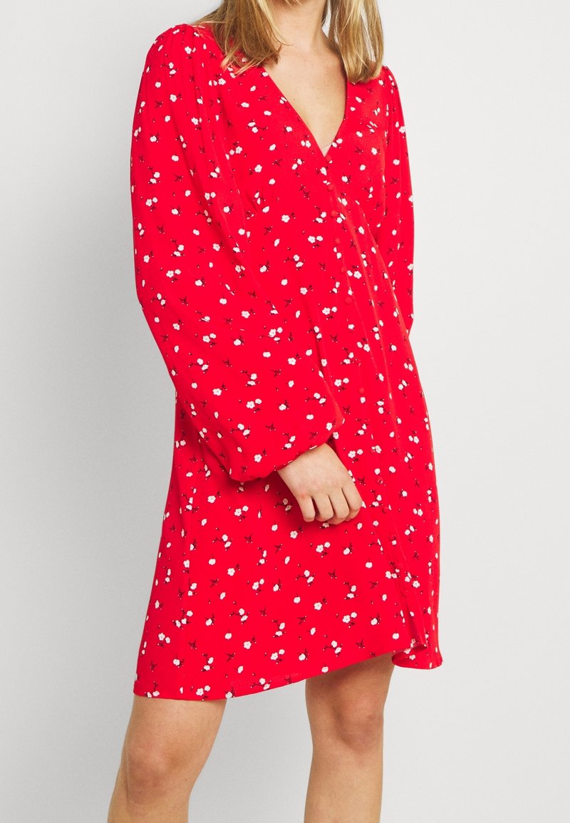 Femme portant une robe rouge avec de longues manches bouffantes et de petits motifs floraux blancs, debout devant un fond clair uni.