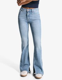Lyseblå flagrende jeans med høj talje, der har forlommer, sømdetaljer og sideslidser ved kanten, parret med hvide sneakers.