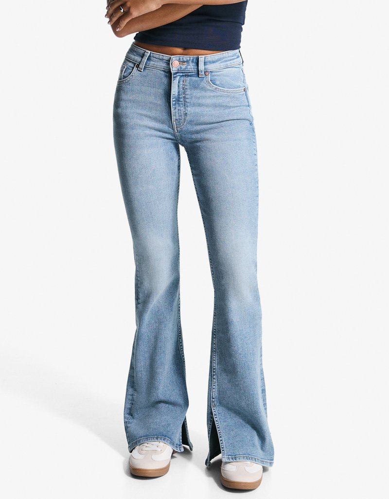 Lyseblå flagrende jeans med høj talje, der har forlommer, sømdetaljer og sideslidser ved kanten, parret med hvide sneakers.