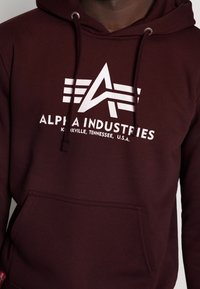 Felpa color maroon con logo bianco di Alpha Industries e testo "Knoxville, Tennessee, U.S.A." sul petto, dotata di tasca frontale e cordini.