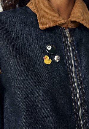 Farkkutakki ruskealla corduroy-kauluksella, jossa on kolme nappia: musta pyöreä nappi, jossa on "8", keltainen kumikala ja hopeinen levy.