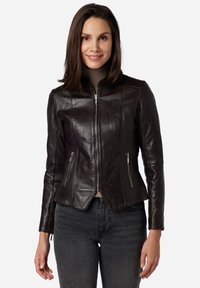 Giacca in pelle marrone con un design aderente, frontale con zip e tasche laterali. Texture liscia e colletto alto. Indossata sopra un top chiaro.
