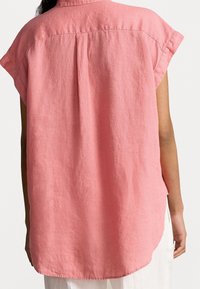 Camicetta di lino rosa con maniche a cappuccio e vestibilità ampia, caratterizzata da una piega sul retro e un orlo arrotondato. Tessuto testurizzato con un leggero effetto lucido.