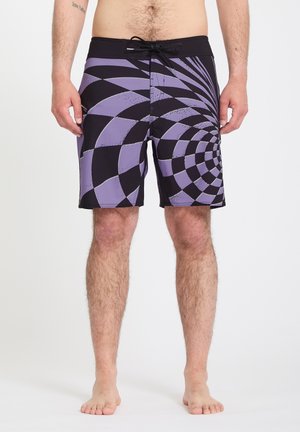 Hombre vistiendo pantalones cortos de baño con un patrón de remolinos a cuadros en negro y morado, de pie descalzo contra un fondo claro y liso.