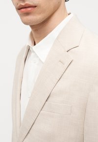 Κοντινό πλάνο ενός ατόμου που φοράει ένα ανοιχτό μπεζ σακάκι με υφή πάνω από ένα λευκό πουκάμισο με κουμπιά, δείχνοντας το πέτο και την τσέπη στο στήθος.