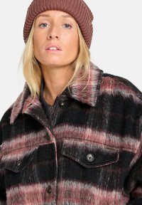 Blonde vrouw in een geribbelde bruine gebreide beanie en een knopen zwart, roze en grijs geruite wollen jas met borstzakken.