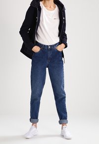 Person wearing rolled-up blue jeans, white sneakers, white T-shirt, and black hoodie with hands in pockets, standing against a plain background.