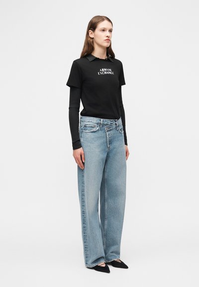 Γυναίκα που φοράει μαύρο μακρυμάνικο στρωματικό τοπ με το κείμενο "Armani Exchange", ανοιχτό μπλε τζιν παντελόνι φαρδιάς γραμμής και μαύρα μυτερά παπούτσια, στέκεται μπροστά σε λευκό φόντο.