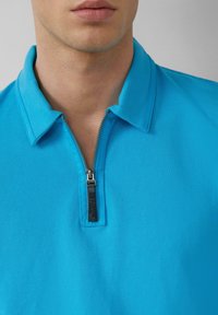Turkusowa koszulka polo z zamkiem błyskawicznym przy kołnierzyku. Wykonana z teksturowanej tkaniny, z małym logo na suwaku dla dodatkowego akcentu.