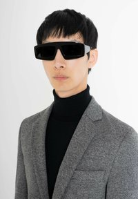 Gafas de sol rectangulares negras con montura gruesa, llevadas por una modelo con un blazer texturizado gris y un cuello de tortuga negro, sobre un fondo blanco.