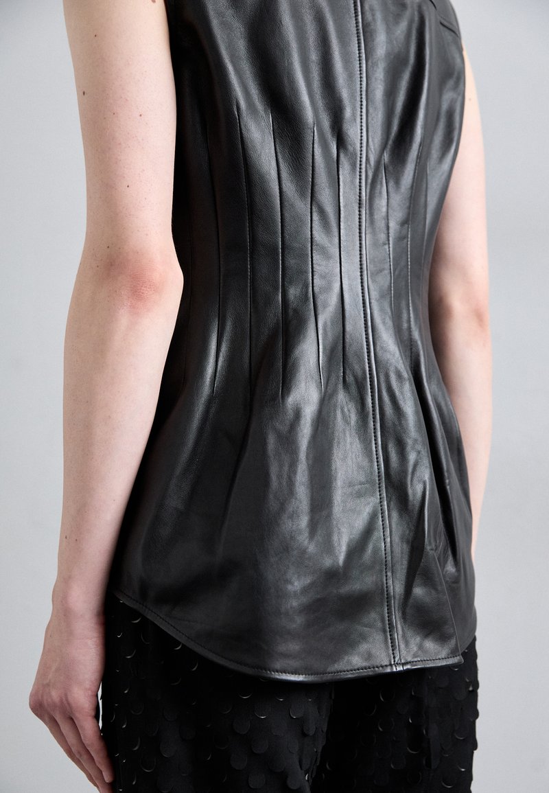 Haut sans manches en cuir noir avec des plis verticaux, ourlet arrondi et fermeture éclair centrale, associé à un pantalon noir texturé avec des accents circulaires.