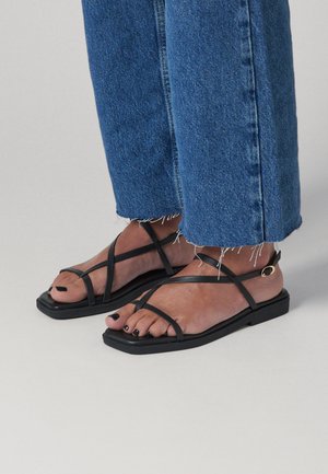 Piedi con sandali neri con cinturini, punte quadrate e smalto nero, abbinati a jeans di denim blu tagliati e sfrangiati.
