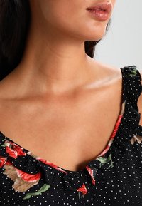 Czarna sukienka w białe kropki, z kwiatowymi akcentami w odcieniach czerwieni i zieleni oraz falbanionym dekoltem. Gładki materiał o dopasowanym kroju.