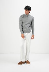Mann in grauem Zip-up-Pullover, weißen Hosen und braunen Slippern steht und schaut seitlich vor einem weißen Hintergrund.