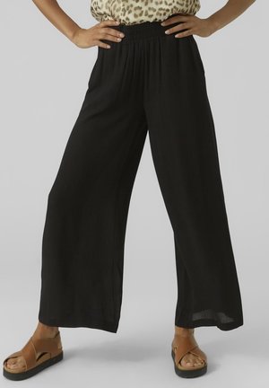 Persona con pantalones anchos negros, top de estampado de leopardo y sandalias marrones con tiras cruzadas, de pie con las manos en las caderas.