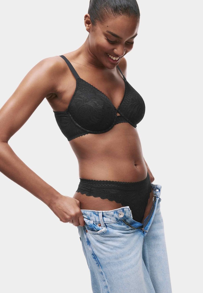 Zwarte kant bh met golvende randen en bijpassende string, gecombineerd met losse lichtblauwe denim jeans, vastgehouden bij de taille.