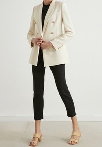Double-breasted cream blazer with gold buttons, tailored fit, soft texture, paired with black cropped pants and beige open-toe heels.