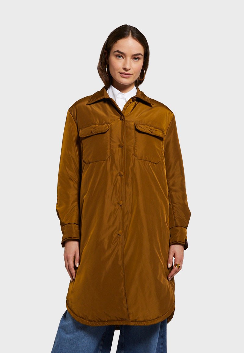 Dlhý, hnedý, lesklý nylonový kabát s gombíkovým zapínaním, dvoma náprsníkmi, špicatým golierom a mierne zakriveným lemom.