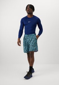 Marineblaue langärmlige Sportshirt, türkisfarbene Shorts mit schwarzem Leopardenmuster und schwarze Sneakers mit gelben Akzenten. Strukturiertes Gewebe insgesamt.