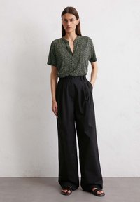 Korte mouw blouse in donkergroen met een verspreid wit patroon, gecombineerd met hoge, zwarte wijde broek en zwarte sandalen.