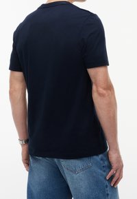 Homme portant un t-shirt bleu marine foncé uni à manches courtes avec un jean en denim bleu clair, vu de dos.