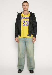 Giacca nera leggera sopra una canotta da basket gialla con dettagli viola e numero 23, abbinata a jeans azzurri a gamba larga e scarpe nere.