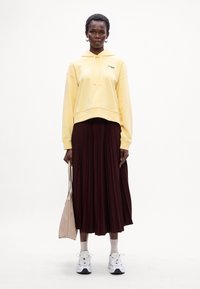 Cropped hoodie gialla con logo, gonna bordeaux plissettata e sneakers bianche. Accessorio: borsa tote beige. Lo sfondo è bianco uniforme.
