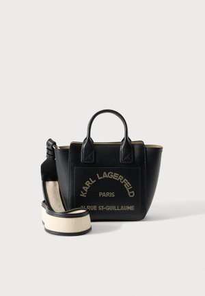 Czarna skórzana torba typu tote z krótkimi uchwytami i odpinanym szerokim beżowym paskiem, z złotym napisem "Karl Lagerfeld Paris" na przedniej kieszeni.