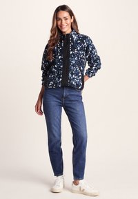 Navy fleece jacket with multicolored abstract pattern, featuring a black zip closure and side pockets, paired with blue denim jeans and white sneakers.