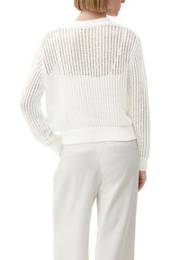 Vitt stickat tröja med ett öppet vävd mönster, ribbade ärmslut och nederkant, med en genomskinlig rygg och avslappnad passform. Bärs med ljusa byxor.