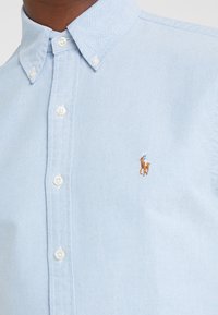 Ljusblå vävd skjorta med krage som knäpps, som har vita knappar och en liten broderad hästlogotyp i brunt och blått på vänster bröst.