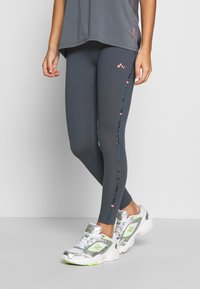 Šedé sportovní legíny s přiléhavým střihem, které mají boční vzor v modrozelených a růžových akcentách. V kombinaci se stříbrnými a neonově zelenými teniskami.