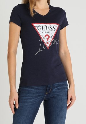 Kvinde iført en tætsiddende marineblå V-hals T-shirt med et rødt og hvidt Guess-logo og blå jeans, stående med afslappede hænder.