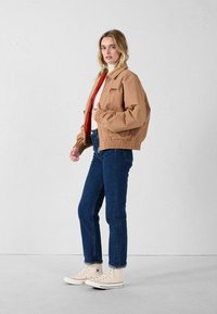 Giacca marrone corta con polsini elasticizzati, indossata sopra una maglia rossa, abbinata a jeans di denim blu e sneakers beige alte. Design semplice, texture liscia.