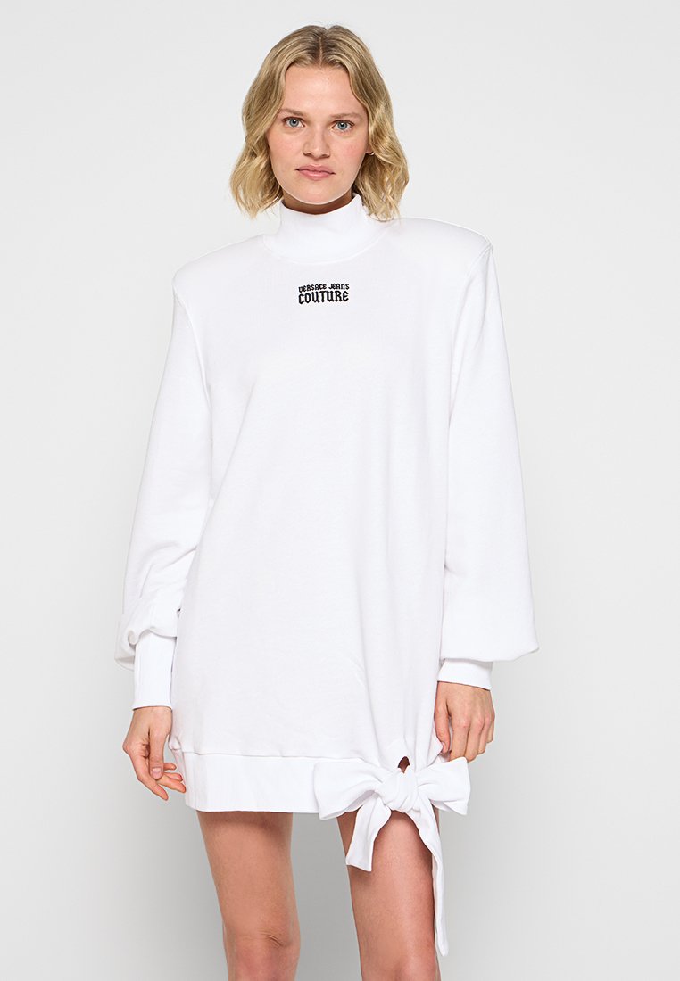 Oversized biały sweter-dress z wysokim dekoltem i kokardką po boku. Posiada czarne logo "Versace Jeans Couture". Miękka tkanina.