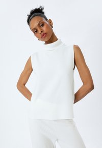 Haut sans manches en col roulé blanc avec une texture côtelée, présentant un col haut et une coupe décontractée. Tissu lisse sans motifs ni accents.