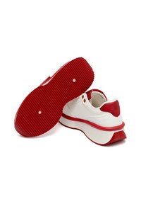 Sneaker bianca con un accentuato tallone rosso testurizzato e suola in gomma rossa con scanalature, progettata con un'ampia intersuola bianca e occhielli metallici.